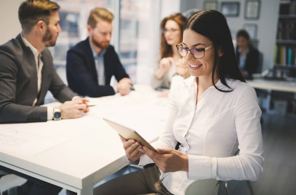 Geschäftsfrau mit Tablet lächelnd bei einem Teammeeting, steht für Teamarbeit und digitale Effizienz bei der Implementierung von Informationssicherheits-Managementsystemen.